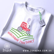Футболка найкращий тракторист. Біла футболка, зображена зверху, з коротким рукавом. На фоні світло-сірий колір. Принт містить червоний трактор, що курить, на зелених полях, з написом 'найкращий тракторист' у червоному кольорі. На футболці також є етикетка.