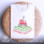 Футболка найкращий тракторист. Біла футболка, складена на плетеній основі, зображена зверху. На фоні світло-сірий колір. Принт містить червоний трактор, що курить, на зелених полях, з написом 'найкращий тракторист' у червоному кольорі.
