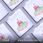 Футболка найкращий тракторист. Біла футболка з коротким рукавом, зображена з фронтального ракурсу на манекені. На фоні світло-сірий колір. Принт містить червоний трактор, що курить, на зелених полях, з написом 'найкращий тракторист' у червоному кольорі.