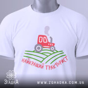 Футболка найкращий тракторист. Біла футболка, зображена зверху, з коротким рукавом. На фоні світло-сірий колір. Принт містить червоний трактор, що курить, на зелених полях, з написом 'найкращий тракторист' у червоному кольорі. На футболці також є етикетка.