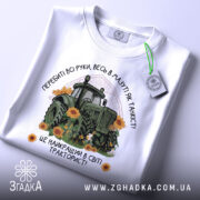 Футболка найкращий в світі тракторист. Біла футболка з принтом, на якому зображений зелений трактор серед соняшників, з етикеткою на комірці. Фон світлий, футболка зображена з верхнього ракурсу.