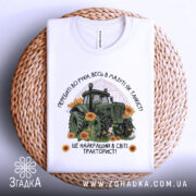 Футболка найкращий в світі тракторист. Біла футболка з принтом, на якому зображений зелений трактор серед соняшників, розкладена на плетеній основі. Фон світлий, футболка зображена зверху.