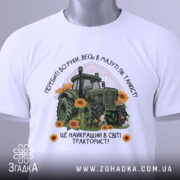 Футболка найкращий в світі тракторист. Біла футболка з принтом, на якому зображений зелений трактор серед соняшників, з етикеткою на комірці. Фон світлий, футболка зображена з верхнього ракурсу.