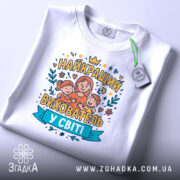 Футболка найкращий вихователь, білого кольору, зображена зверху, на фоні світло-сірого кольору, принт з малюнком жінки з двома дітьми та написом 'НАЙКРАЩИЙ ВИХОВАТЕЛЬ У СВІТІ', на футболці є етикетка з зеленим шнурком.