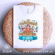 Футболка найкращий вихователь, білого кольору, зображена зверху на плетеній основі, фон світло-сірий, принт містить малюнок жінки з двома дітьми та написом 'НАЙКРАЩИЙ ВИХОВАТЕЛЬ У СВІТІ' у яскравих кольорах.