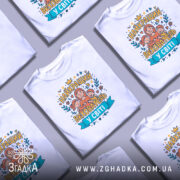 Футболка найкращий вихователь, білого кольору, зображена на манекені з правого боку, фон світло-сірий, принт містить малюнок жінки з двома дітьми та написом 'НАЙКРАЩИЙ ВИХОВАТЕЛЬ У СВІТІ' у яскравих кольорах.