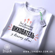Футболка найкращий вихователь, біла, складена на столі, фон світло-сірий, принт чорного кольору з оранжевими стрілками, на ярлику зелена стрічка.