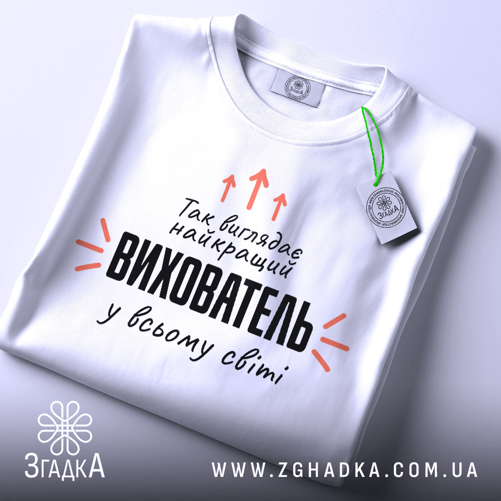 Футболка найкращий вихователь, біла, складена на столі, фон світло-сірий, принт чорного кольору з оранжевими стрілками, на ярлику зелена стрічка.