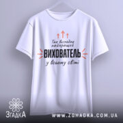Футболка найкращий вихователь, біла, підвішена на вішалці, фон світло-сірий, принт чорного кольору з оранжевими стрілками та текстом.