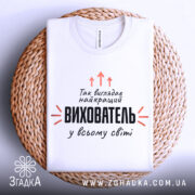 Футболка найкращий вихователь, біла, складена на плетеній кошику, фон світло-сірий, принт чорного кольору з оранжевими стрілками та текстом.