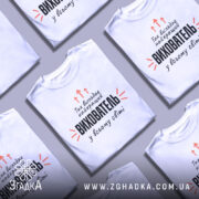 Футболка найкращий вихователь, біла, зображена на манекені, фон світло-сірий, принт чорного кольору з оранжевими стрілками та текстом.