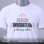 Футболка найкращий вихователь, біла, складена на столі, фон світло-сірий, принт чорного кольору з оранжевими стрілками, на ярлику зелена стрічка.