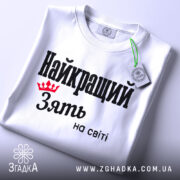 Футболка найкращий зять, біла, складена на поверхні, фон світло-сірий, принт чорного кольору з червоною короною та текстом 'Найкращий зять на світі', з етикеткою на шнурку.