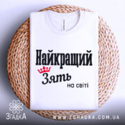 Футболка найкращий зять, біла, складена на плетеній кошику, фон світло-сірий, принт чорного кольору з червоною короною та текстом 'Найкращий зять на світі'.