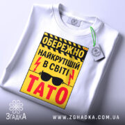 Футболка найкрутіший тато, біла, зображена зверху, фон світлий, принт жовтий з чорними і червоними елементами, текст з попередженням і написом 'ТАТО', на футболці є етикетка з зеленим шнурком.