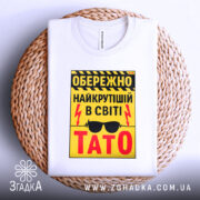 Футболка найкрутіший тато, біла, зображена на плоскій поверхні, фон світлий, принт жовтий з чорними і червоними елементами, текст з попередженням і написом 'ТАТО', розташована на плетеній основі.
