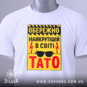 Футболка найкрутіший тато, біла, зображена зверху, фон світлий, принт жовтий з чорними і червоними елементами, текст з попередженням і написом 'ТАТО', на футболці є етикетка з зеленим шнурком.