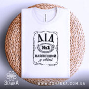 Футболка найліпший дід, білого кольору, зображена зверху на плоскій поверхні, фон світло-сірий, принт чорного кольору з написом 'ДІД №1 НАЙЛІПШИЙ у світі', футболка розташована на плетеній основі.