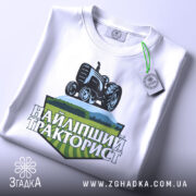 Футболка найліпший тракторист. Біла футболка, складена на столі, з видимим принтом. На фоні світло-сірий колір. Принт з трактором та написом 'НАЙЛІПШИЙ ТРАКТОРИСТ' у зелених та чорних тонах, з етикеткою на зеленій стрічці.