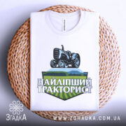 Футболка найліпший тракторист. Біла футболка, складена на плетеній кошику, з видимим принтом. На фоні світло-сірий колір. Принт з трактором та написом 'НАЙЛІПШИЙ ТРАКТОРИСТ' у зелених та чорних тонах.