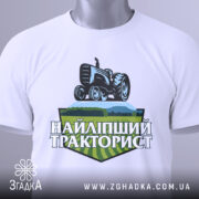 Футболка найліпший тракторист. Біла футболка, складена на столі, з видимим принтом. На фоні світло-сірий колір. Принт з трактором та написом 'НАЙЛІПШИЙ ТРАКТОРИСТ' у зелених та чорних тонах, з етикеткою на зеленій стрічці.