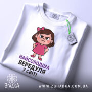 Футболка найсолодша вередуля, білого кольору, зображена зверху, на ній видно етикетку з зеленим шнурком, фон світлий, принт з малюнком дівчинки у рожевій сукні та написом 'НАЙСОЛОДША ВЕРЕДУЛЯ У СВІТІ'.