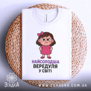 Футболка найсолодша вередуля, білого кольору, зображена зверху на плетеній основі, фон світлий, принт з малюнком дівчинки у рожевій сукні та написом 'НАЙСОЛОДША ВЕРЕДУЛЯ У СВІТІ'.