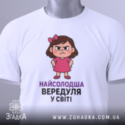 Футболка найсолодша вередуля, білого кольору, зображена зверху, на ній видно етикетку з зеленим шнурком, фон світлий, принт з малюнком дівчинки у рожевій сукні та написом 'НАЙСОЛОДША ВЕРЕДУЛЯ У СВІТІ'.