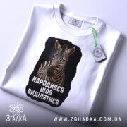 Футболка народився щоб виділятися, складена біла футболка з принтом зебри, з ракурсу зверху, на світлому фоні, з етикеткою, що звисає.