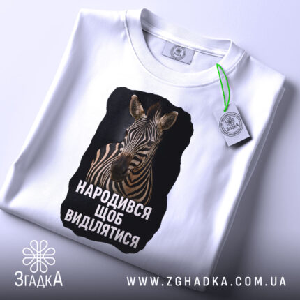 Футболка народився щоб виділятися, складена біла футболка з принтом зебри, з ракурсу зверху, на світлому фоні, з етикеткою, що звисає.