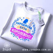 Футболка народжена бути королевою, білого кольору, складена на плоскій поверхні, фон світлий, принт з короною та написом в яскравих кольорах: синьому, рожевому та золотому, з видимою етикеткою.