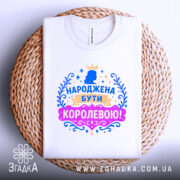 Футболка народжена бути королевою, білого кольору, складена на круглій плетеній основі, фон світлий, принт з короною та написом в яскравих кольорах: синьому, рожевому та золотому.