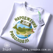 Футболка народжений для риболовлі, білого кольору, зображена зверху, на ній видно етикетку, фон світло-сірий, принт з рибою та текстом в зелених відтінках.