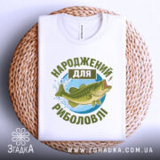 Футболка народжений для риболовлі, білого кольору, зображена зверху на плетеному кошику, фон світло-сірий, принт з рибою та текстом в зелених відтінках.