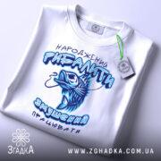 Футболка народжений рибалити, білого кольору, складена на поверхні, фон світлий, принт з рибою та текстом у блакитних відтінках, на футболці є етикетка з зеленим шнурком.