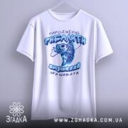 Футболка народжений рибалити, білого кольору, підвішена на вішалці, фон світлий, принт з рибою та текстом у блакитних тонах, видно округлі краї рукавів.