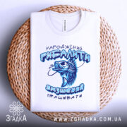 Футболка народжений рибалити, білого кольору, складена на плетеній кошику, фон світлий, принт з рибою та текстом у блакитних відтінках, видно округлий виріз горловини.