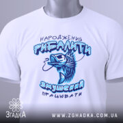 Футболка народжений рибалити, білого кольору, складена на поверхні, фон світлий, принт з рибою та текстом у блакитних відтінках, на футболці є етикетка з зеленим шнурком.