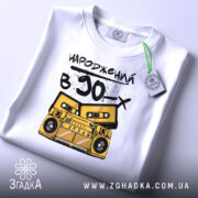 Футболка народжений в 90, складена на столі, фон світлий, принт з жовтим магнітофоном і касетами, текст чорний, на футболці є етикетка з зеленим шнурком.