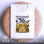 Футболка народжений в 90, складена на плетеній кошику, фон світлий, принт з жовтим магнітофоном і касетами, текст чорний, виглядає акуратно.