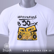 Футболка народжений в 90, складена на столі, фон світлий, принт з жовтим магнітофоном і касетами, текст чорний, на футболці є етикетка з зеленим шнурком.