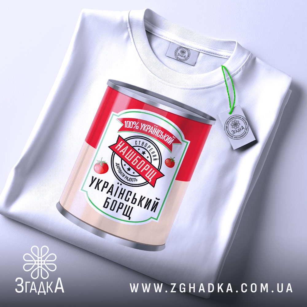 Футболка наш борщ. Біла футболка, складена на поверхні, з принтом банки борщу. Принт яскравий, з червоними помідорами та написом '100% український'. На футболці є етикетка з зеленим шнурком.