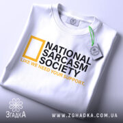 Футболка National Sarcasm Society білого кольору, складена на столі, фон світло-сірий, принт містить чорний текст з жовтим логотипом National Geographic та жовтим написом 'LIKE WE NEED YOUR SUPPORT', на футболці є етикетка з зеленим шнурком.