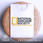 Футболка National Sarcasm Society білого кольору, складена на плетеній кошику, фон світло-сірий, принт містить чорний текст з жовтим логотипом National Geographic та жовтим написом 'LIKE WE NEED YOUR SUPPORT'.