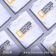 Футболка National Sarcasm Society білого кольору, зображена на манекені з блискучою поверхнею, фон світло-сірий, принт містить чорний текст з жовтим логотипом National Geographic та жовтим написом 'LIKE WE NEED YOUR SUPPORT'.