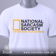 Футболка National Sarcasm Society білого кольору, складена на столі, фон світло-сірий, принт містить чорний текст з жовтим логотипом National Geographic та жовтим написом 'LIKE WE NEED YOUR SUPPORT', на футболці є етикетка з зеленим шнурком.