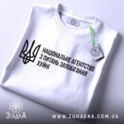Футболка національне агентство, біла, складена на поверхні, фон світлий, принт чорного кольору з текстом та тризубом, з етикеткою, що звисає.