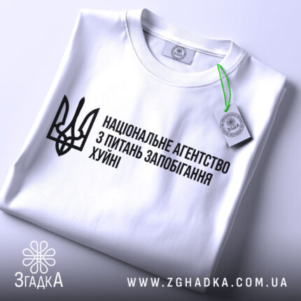 Футболка національне агентство, біла, складена на поверхні, фон світлий, принт чорного кольору з текстом та тризубом, з етикеткою, що звисає.