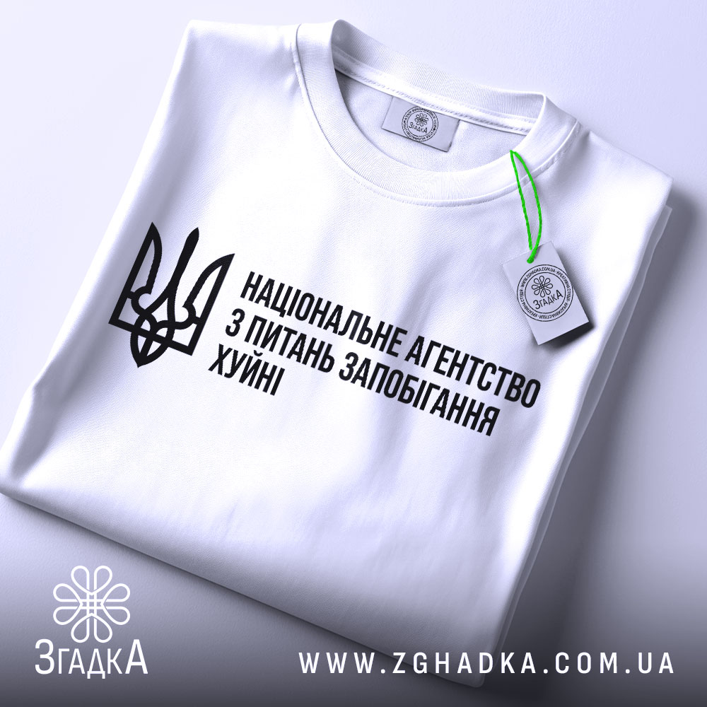 Футболка національне агентство, біла, складена на поверхні, фон світлий, принт чорного кольору з текстом та тризубом, з етикеткою, що звисає.