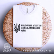 Футболка національне агентство, біла, складена на плетеній кошику, фон нейтральний, принт чорного кольору з текстом та тризубом.